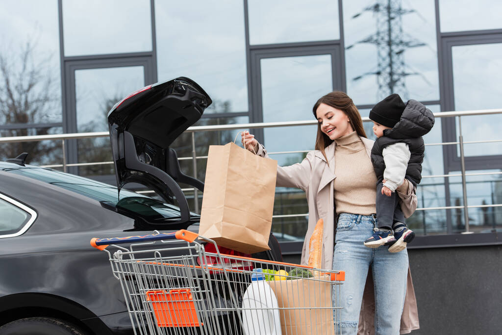 sonriente mujer sosteniendo hijo y bolsa de compras cerca de coche con baúl abierto - Foto, imagen