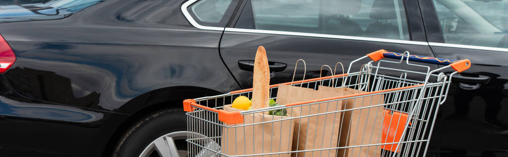 nákupní vozík s čerstvým nákupem v blízkosti černého auta, banner - Fotografie, Obrázek