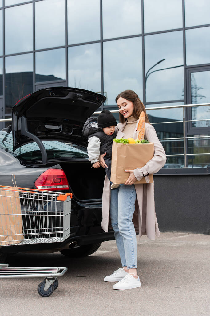 Junge Frau mit Kleinkind kauft nahe Auto mit offenem Kofferraum - Foto, Bild