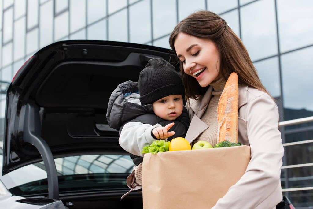 niño apuntando a la comida fresca en la bolsa de compras cerca de la madre feliz al aire libre - Foto, Imagen