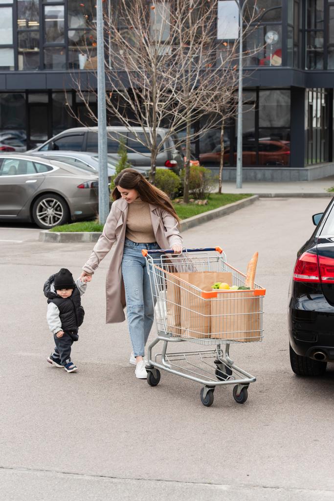 young woman holding hand of child while walking with shopping cart on car parking - Photo, Image
