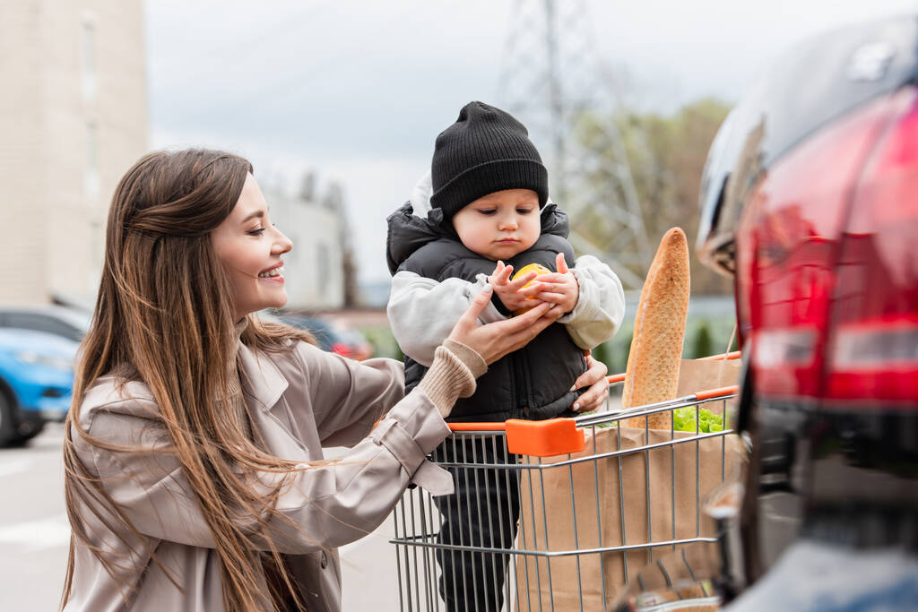 Lächelnde Mutter schenkt Kleinkind-Sohn Orange im Einkaufswagen - Foto, Bild