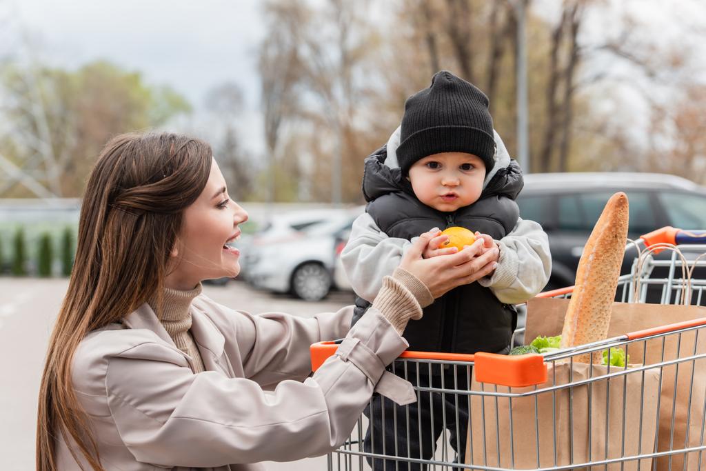happy woman looking at baby boy holding fresh orange while sitting in shopping cart - Photo, Image