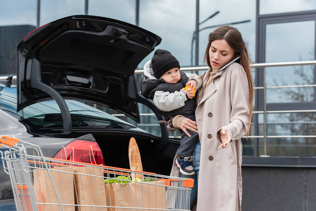 Junge Frau mit Sohn in der Hand telefoniert in der Nähe von Auto und Einkaufswagen mit Smartphone - Foto, Bild