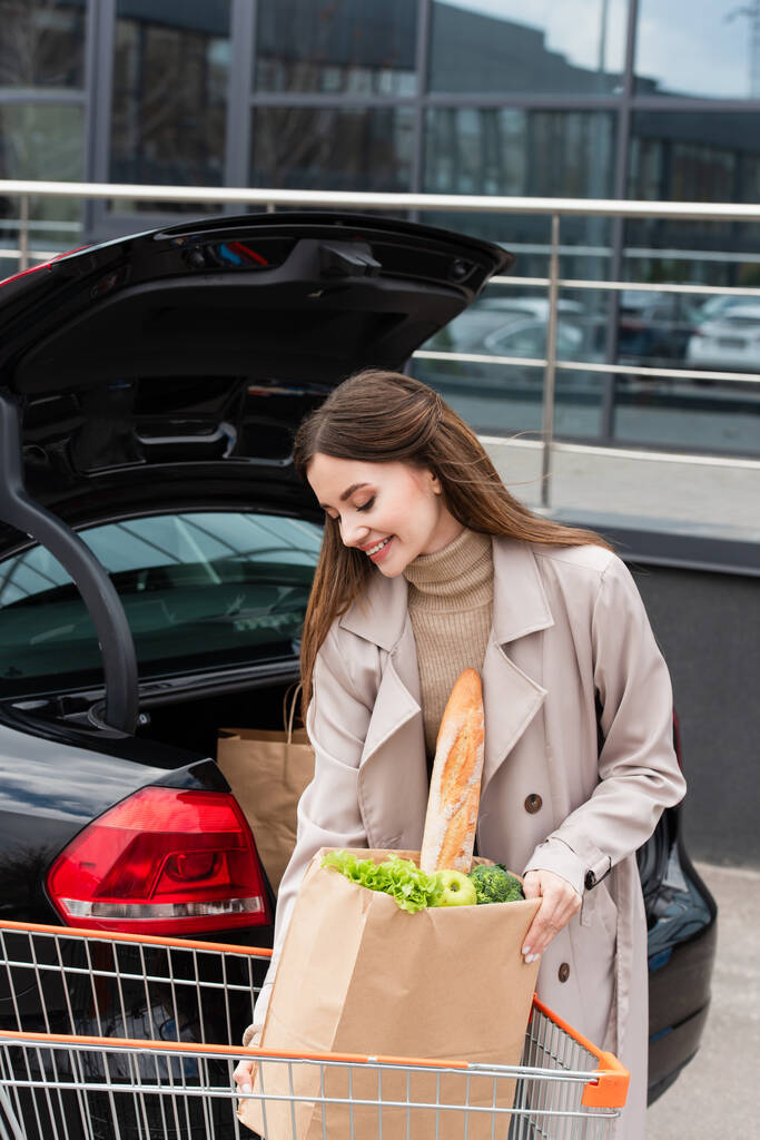pretty, smiling woman holding shopping bag near open car trunk - Photo, Image