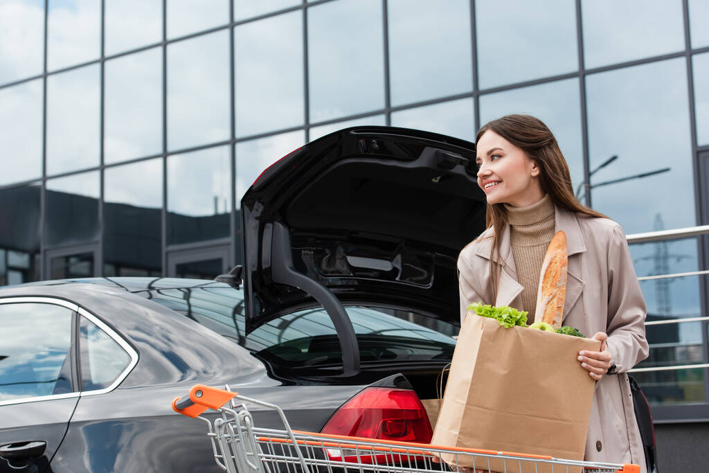szczęśliwa kobieta trzyma torbę na zakupy w pobliżu samochodu z otwartym bagażnikiem  - Zdjęcie, obraz