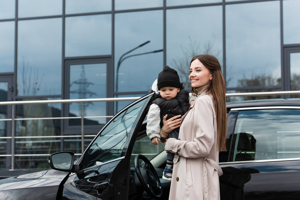 радостная женщина смотрит в сторону, стоя с малышом сыном возле автомобиля - Фото, изображение