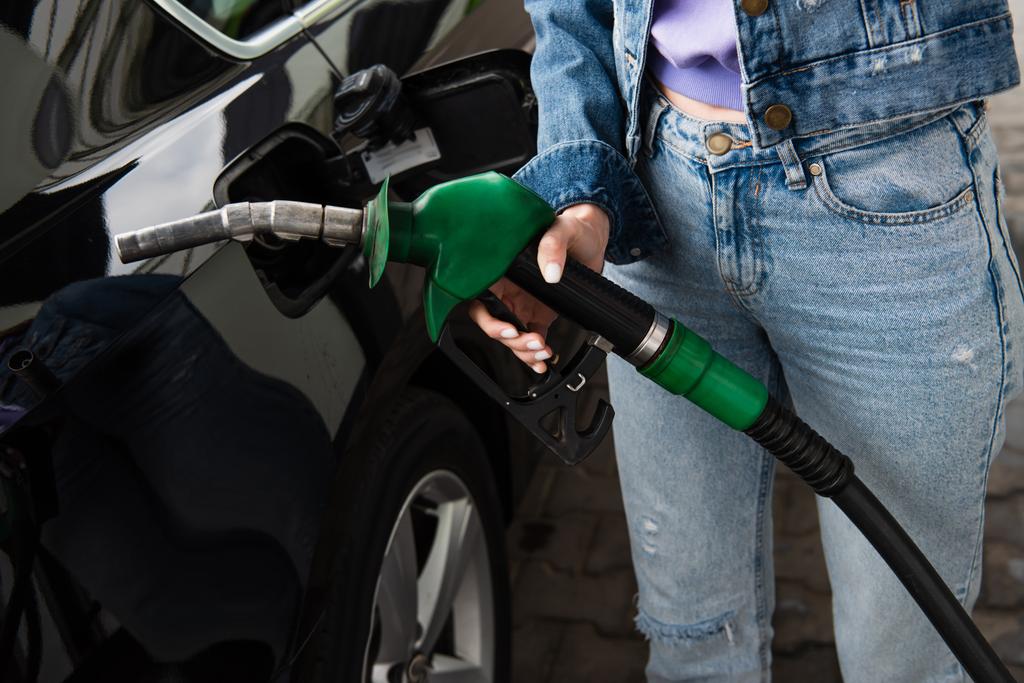Benzin istasyonunda, arabanın yanında elinde yakıt tabancası olan kot pantolonlu bir kadın görülüyor. - Fotoğraf, Görsel