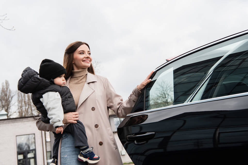 довольная женщина смотрит в сторону, держа маленького сына возле машины на открытом воздухе - Фото, изображение