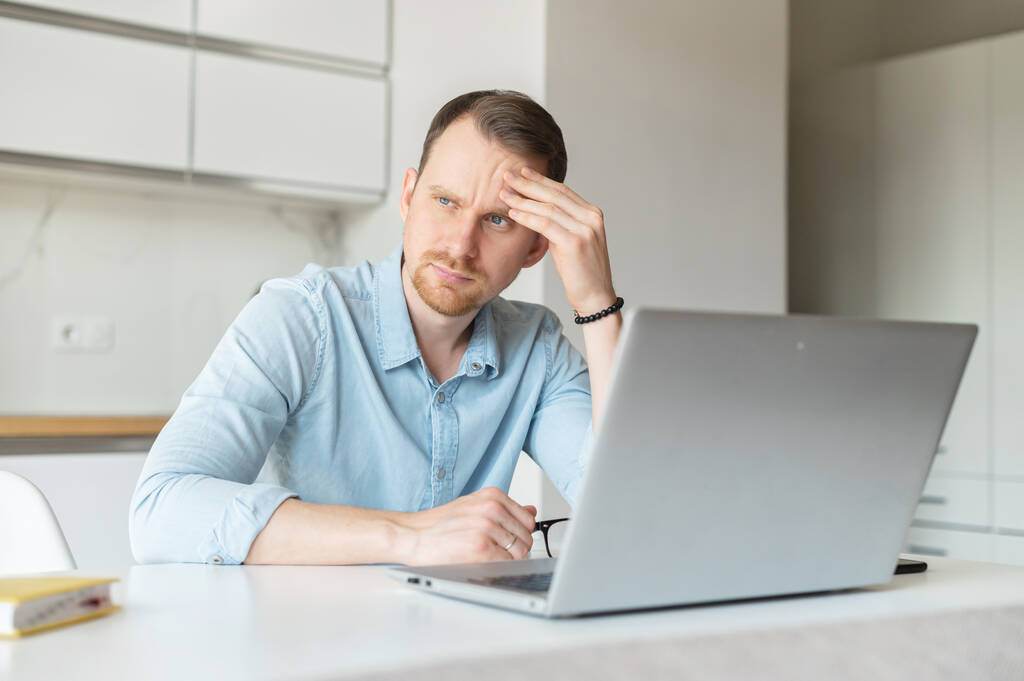 Aggódó fiatalember használja laptop számítógép keresni az állást, munkanélküli fickó ül a konyhában otthon elgondolkodott a jövő, töprengő és ideges férfi álláskereső - Fotó, kép