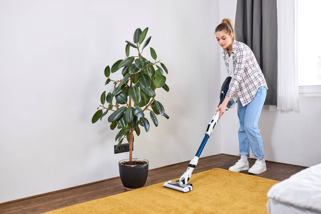 Caucasian young woman wearing casual clothes cleaning house floor in living room using modern vacuum cleaner - Photo, Image