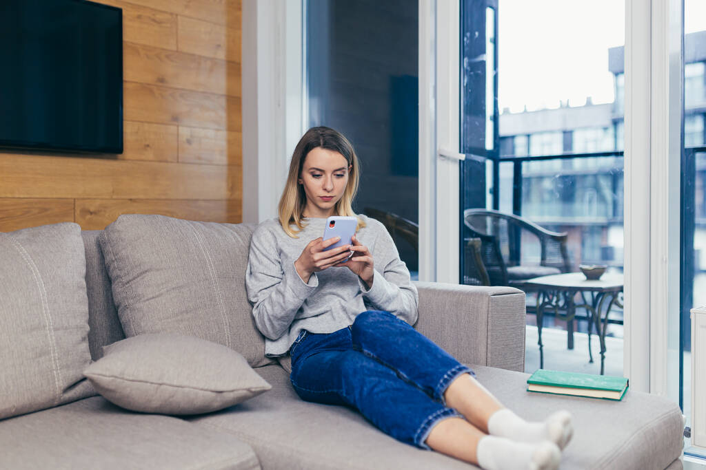 Junge schöne Frau sitzt zu Hause auf der Couch und benutzt ein Mobiltelefon, um sich von der Arbeit auszuruhen - Foto, Bild