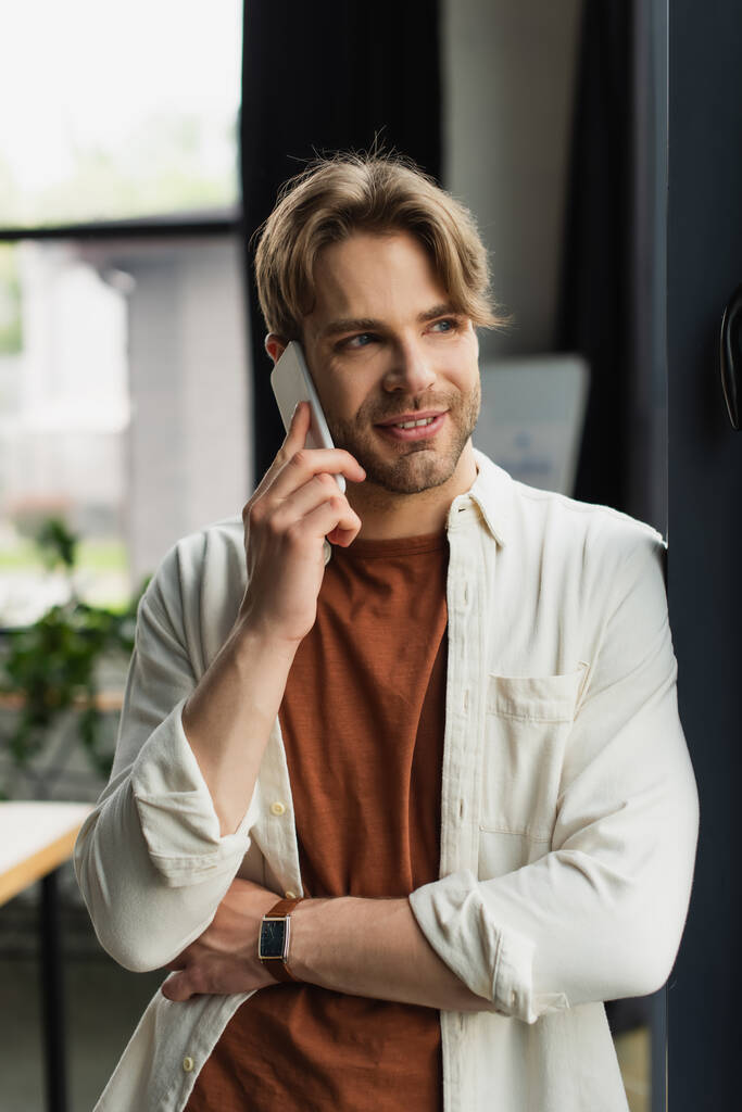 улыбающийся молодой человек в бежевой рубашке разговаривает по мобильному телефону в современном открытом пространстве - Фото, изображение