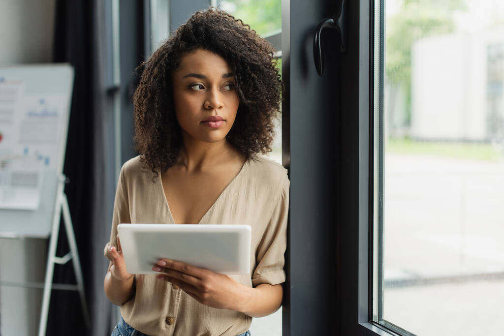 donna afroamericana premurosa in piedi vicino alla finestra con tablet digitale in mano in ufficio moderno - Foto, immagini