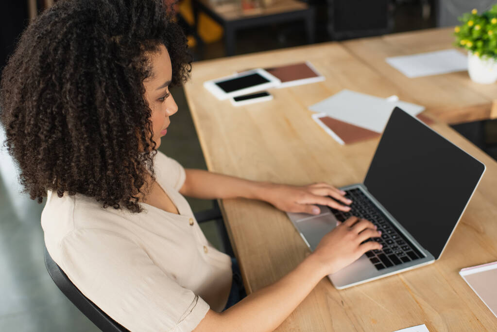 vista de ángulo alto de la mujer afroamericana joven seria escribiendo en el ordenador portátil en la oficina moderna - Foto, imagen