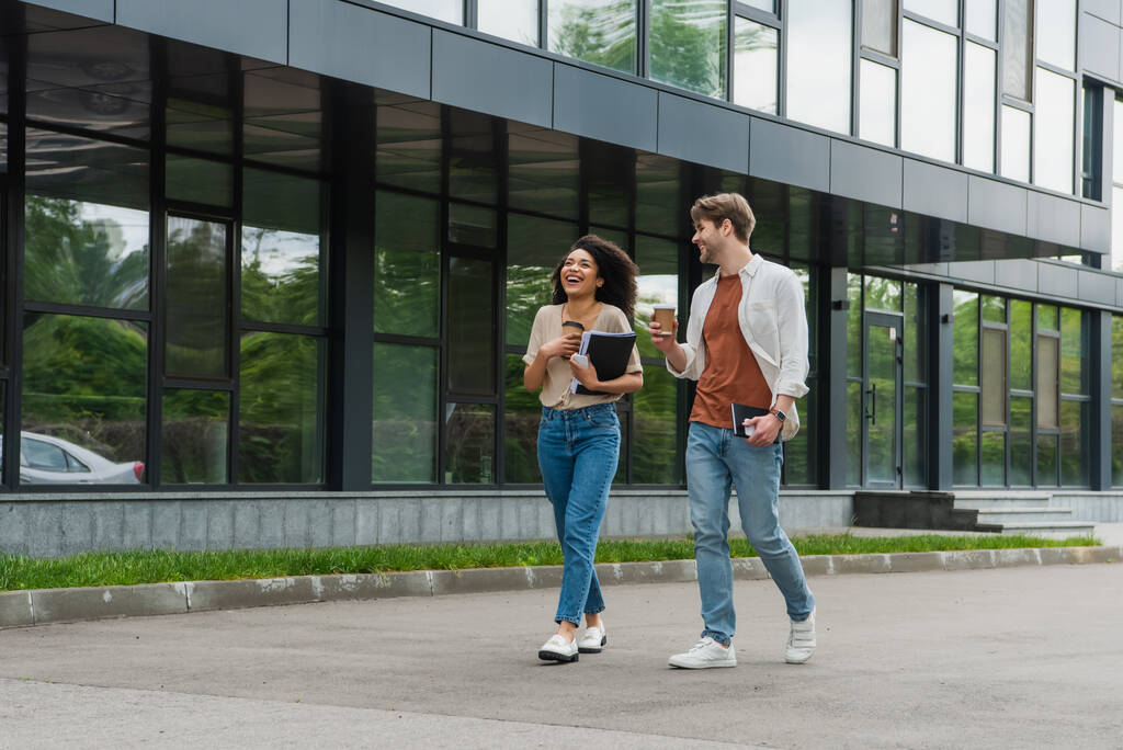 позитивна молода міжрасова пара з паперовими чашками кави та мобільними телефонами в руках, що йдуть по вулиці біля сучасної будівлі
 - Фото, зображення