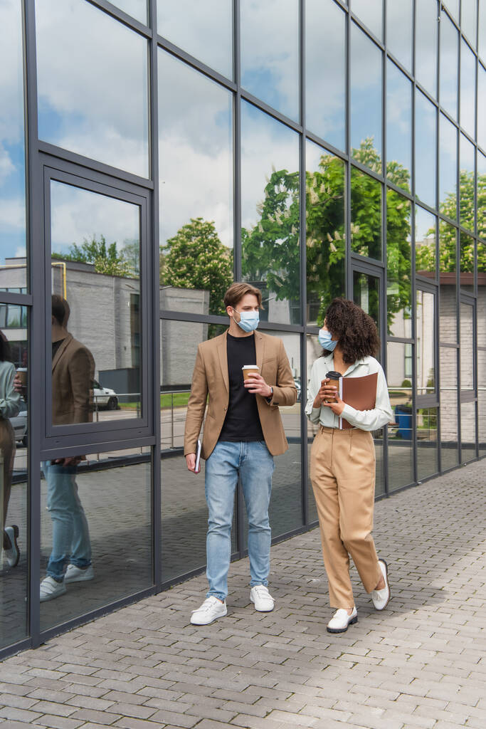 jóvenes colegas interracial en máscaras médicas caminando con tazas de papel de café cerca de edificio moderno - Foto, imagen