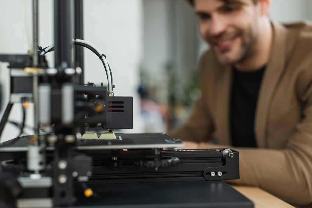 3D принтер, производящий пластиковую модель, рядом с размытым молодым улыбающимся человеком в современном офисе - Фото, изображение
