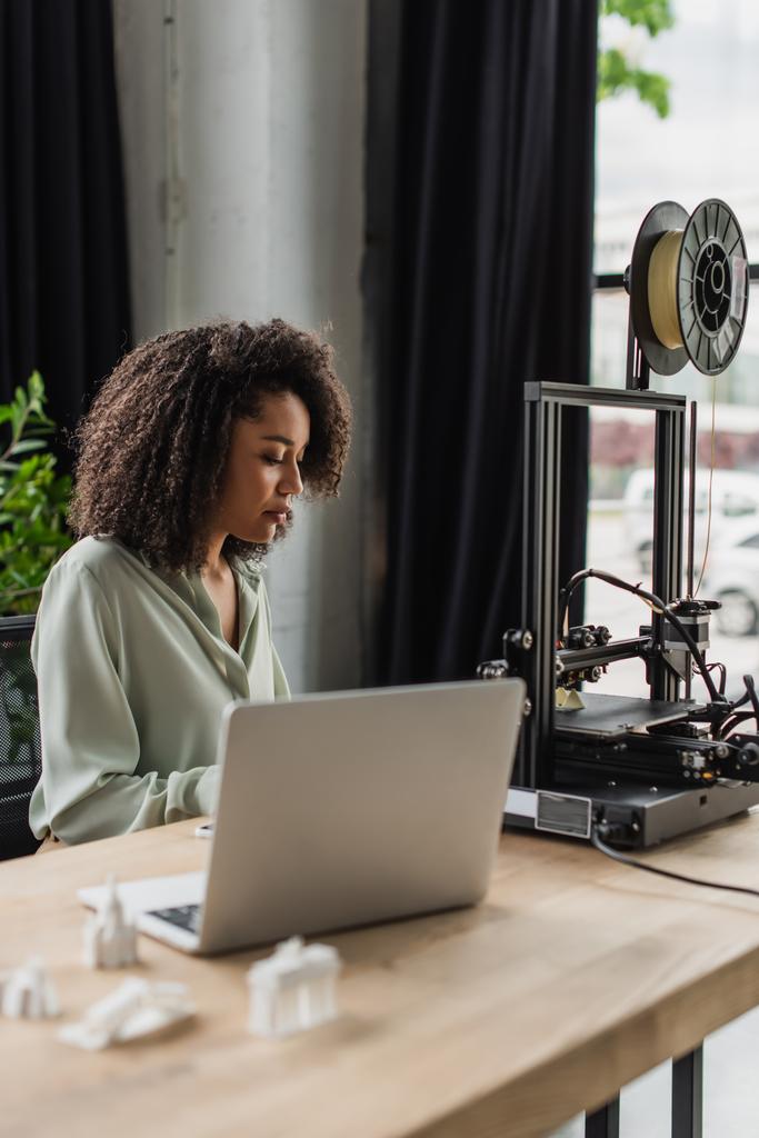 fiatal afro-amerikai tervező ül közelében 3D nyomtató és laptop modern nyílt térben - Fotó, kép