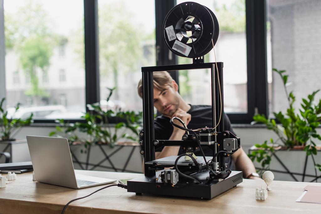 young designer looking at 3D printer on table with laptop in office - Photo, Image