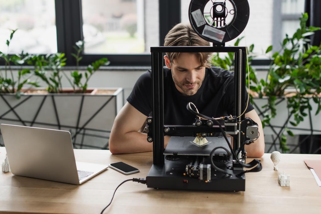 enfocado joven diseñador sentado cerca de la computadora portátil y mirando a la impresora 3D en la oficina moderna - Foto, imagen