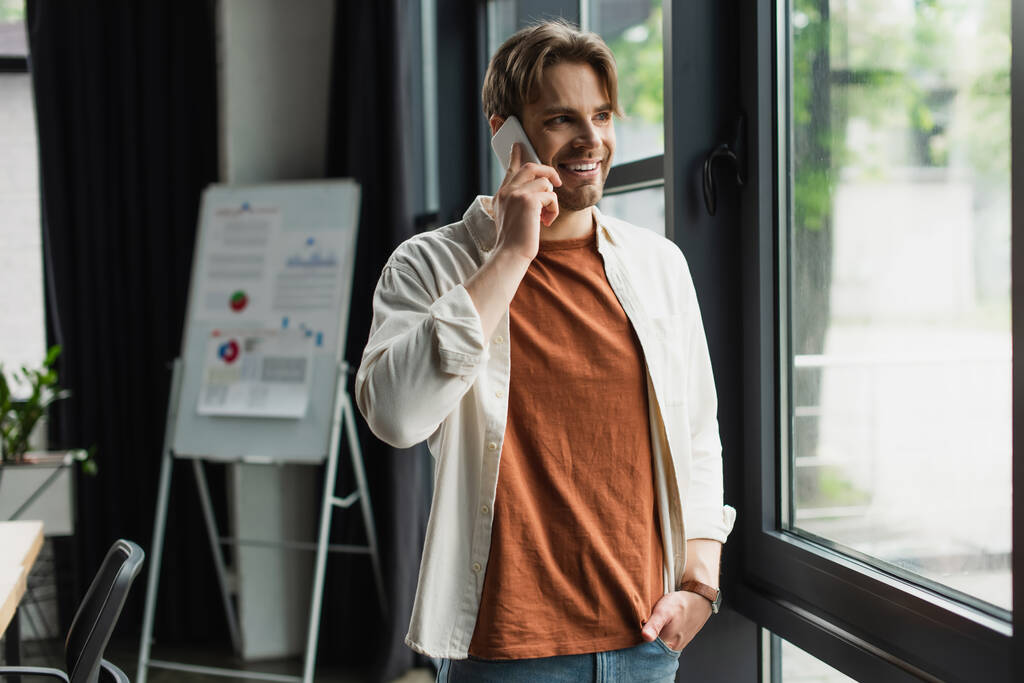позитивный молодой человек разговаривает по мобильному телефону рядом с флип-чартом с графиками и графиками в современном офисе - Фото, изображение