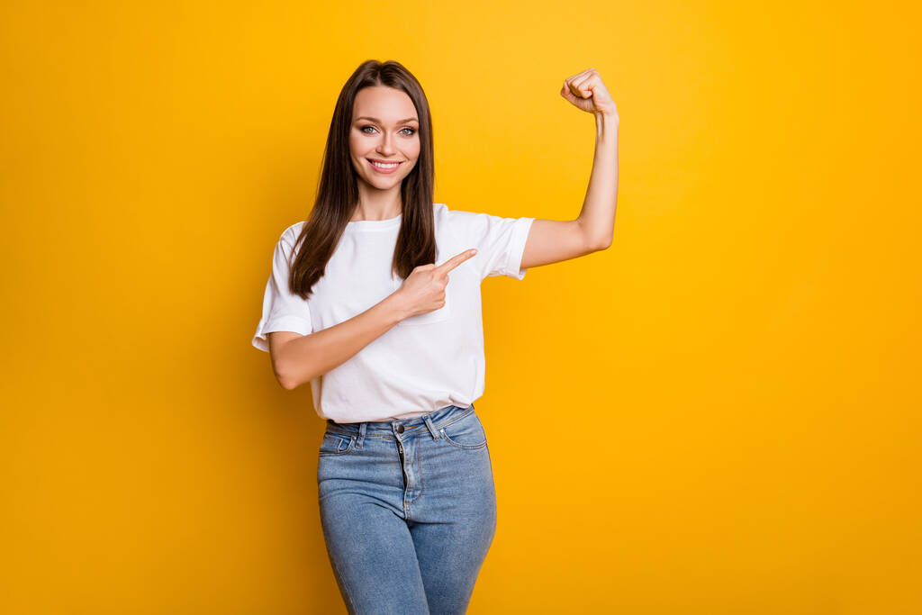 Retrato fotográfico de una chica fuerte señalando con el dedo los bíceps aislados sobre un fondo de color amarillo vivo - Foto, imagen