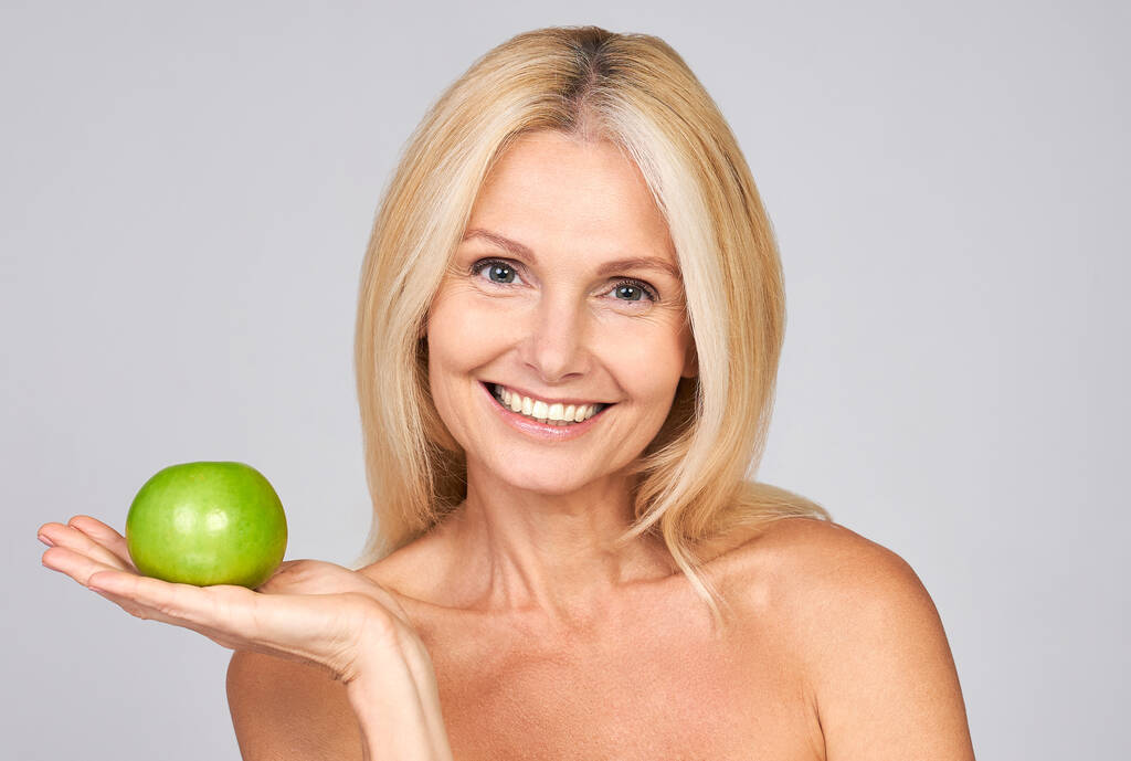 Portrait of a beautiful elderly woman holding an apple, smiling, isolated on gray background. Happy Woman Holding Granny Smith Apple. Dental concept. Copy space. - Photo, Image