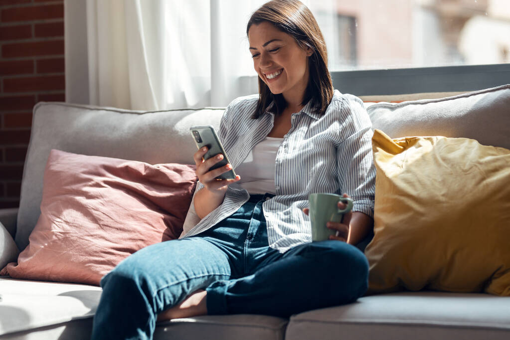 Fotografía de una mujer sonriente bastante joven usando un teléfono móvil sentado en un sofá en casa. - Foto, imagen
