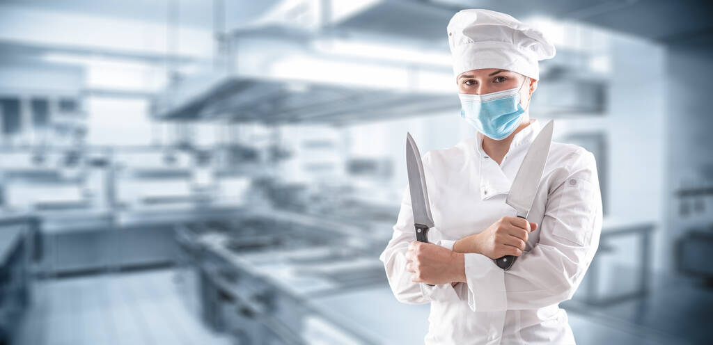 Panoramiczny sztandar szefa kuchni z maską z powodu pandemii covid-19 stojącej ze skrzyżowanymi rękami i trzymającej dwa ostre noże przed kuchnią restauracji. - Zdjęcie, obraz