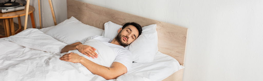 Νεαρός άνδρας με λευκό μπλουζάκι κοιμάται στο κρεβάτι, πανό  - Φωτογραφία, εικόνα