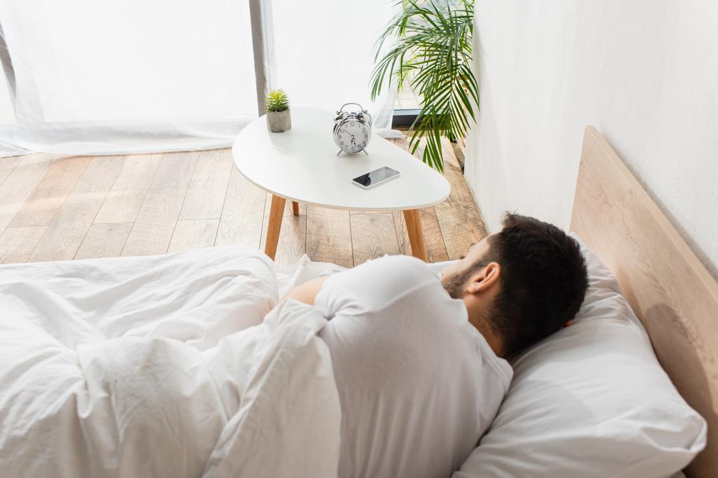 Achteraanzicht van smartphone en wekker op tafel naast man slapend op bed  - Foto, afbeelding