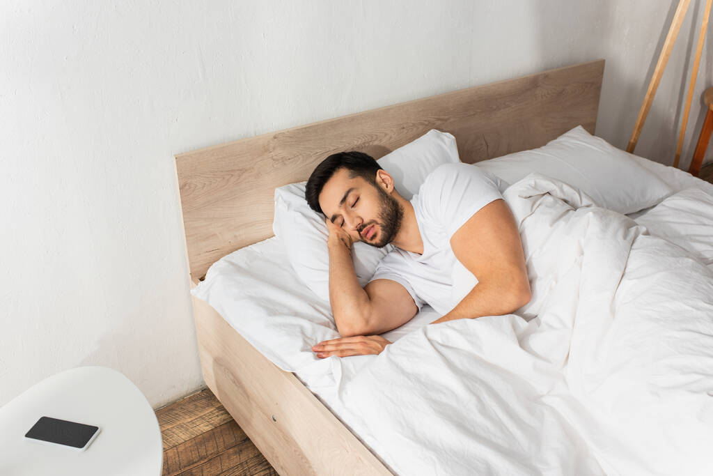 Γενειοφόρος άνθρωπος κοιμάται στο κρεβάτι κοντά smartphone με λευκή οθόνη στο κομοδίνο - Φωτογραφία, εικόνα