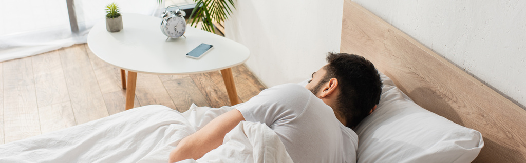 Achteraanzicht van de man die op bed slaapt in de buurt van mobiele telefoon op nachtkastje, banner  - Foto, afbeelding