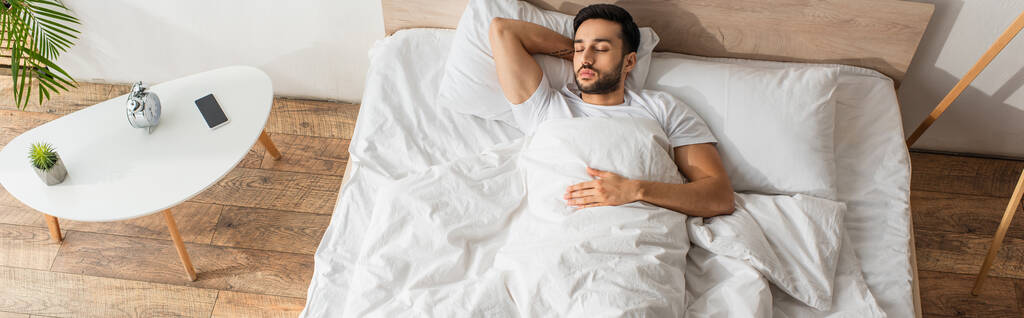 Вид сверху на молодого человека, спящего на кровати возле будильника и мобильного телефона, баннер  - Фото, изображение