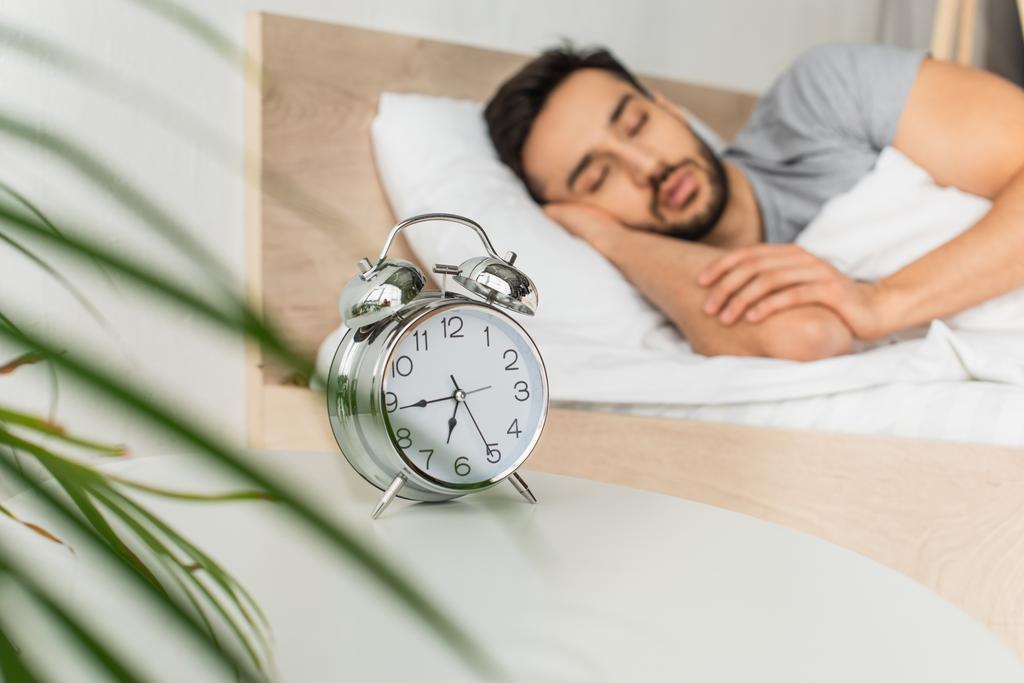 Reloj despertador cerca de la planta borrosa y el hombre durmiendo en la cama  - Foto, Imagen