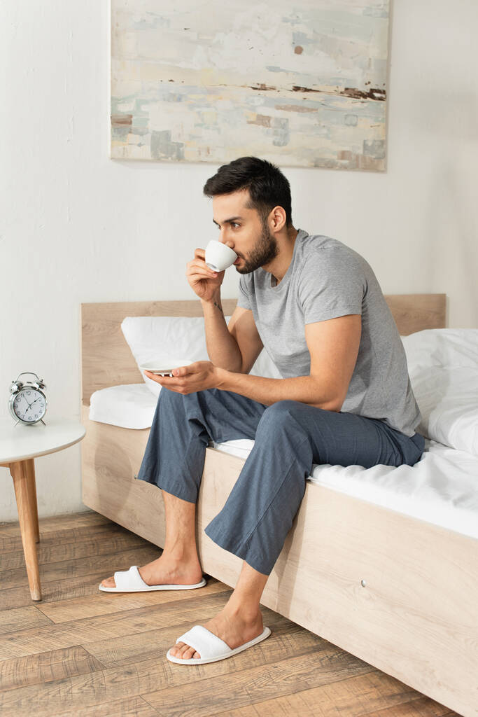 Szakállas férfi pizsamában kávét iszik otthon az ágyon.  - Fotó, kép
