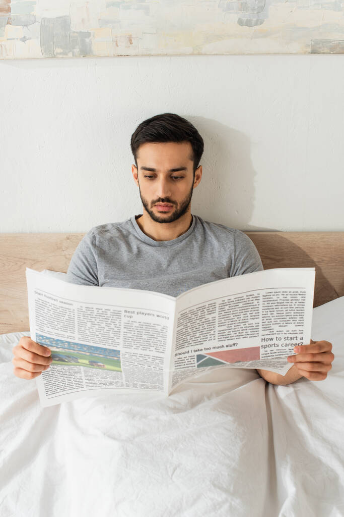 Jongeman in grijs t-shirt leest krant op bed  - Foto, afbeelding
