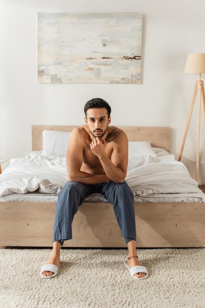 Uomo senza maglietta in pantofole e pantaloni guardando la fotocamera sul letto  - Foto, immagini