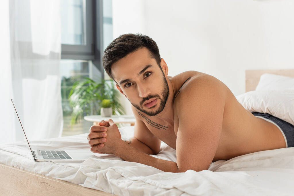 Мужчина без рубашки смотрит в камеру возле ноутбука на кровати дома  - Фото, изображение