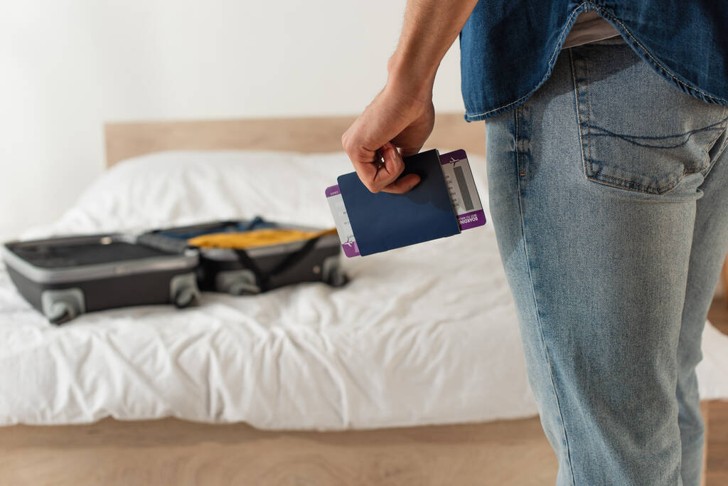 Vista cortada do homem que possui passaporte e bilhete aéreo perto da mala embaçada na cama  - Foto, Imagem
