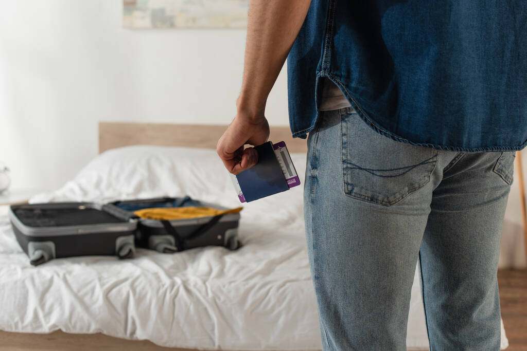 Cropped view of man with passport and boarding pass standing near blurred suitcase in bedroom  - Photo, Image