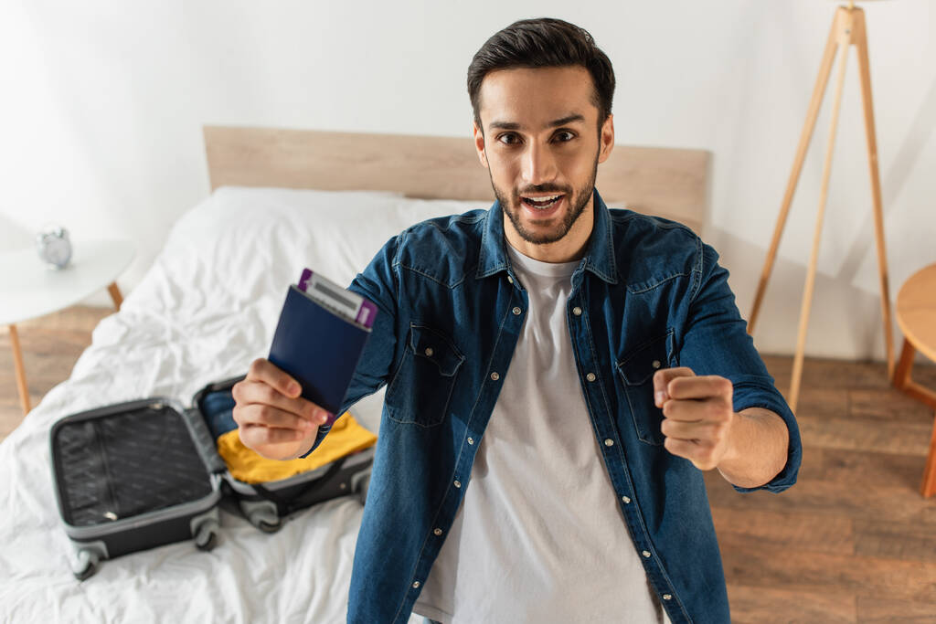 Homme excité tenant un passeport et montrant un geste oui près de la valise floue sur le lit  - Photo, image