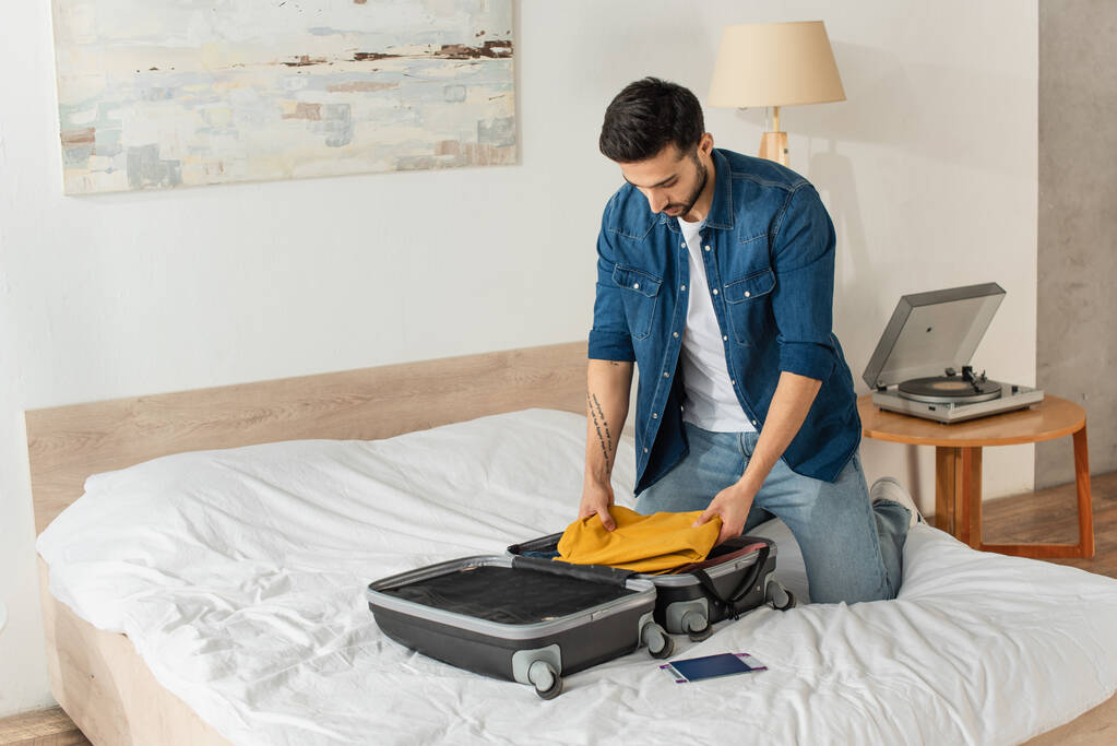 Γενειοφόρος άντρας που βάζει ρούχα σε βαλίτσα κοντά στο διαβατήριο στο κρεβάτι  - Φωτογραφία, εικόνα