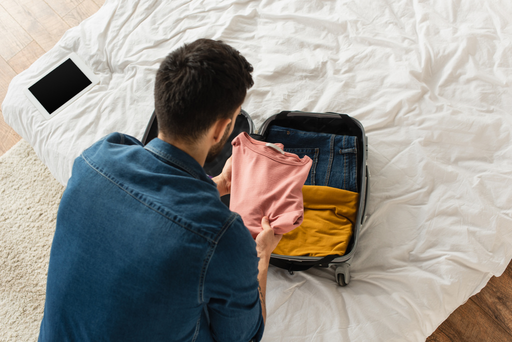 Yleisnäkymä miehestä, joka laittaa vaatteita matkatavaroihin lähellä digitaalista tablettia tyhjällä näytöllä sängyssä  - Valokuva, kuva