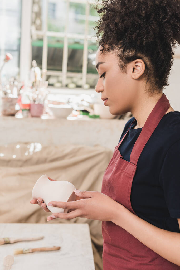 vážná mladá africká americká žena při pohledu na hliněný hrnec v rukou v keramice - Fotografie, Obrázek