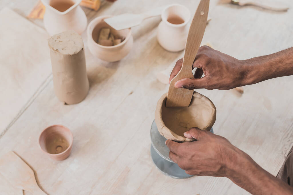 vista parziale del giovane uomo afroamericano che scolpisce vaso di argilla con spatola sul tavolo con attrezzatura in ceramica  - Foto, immagini