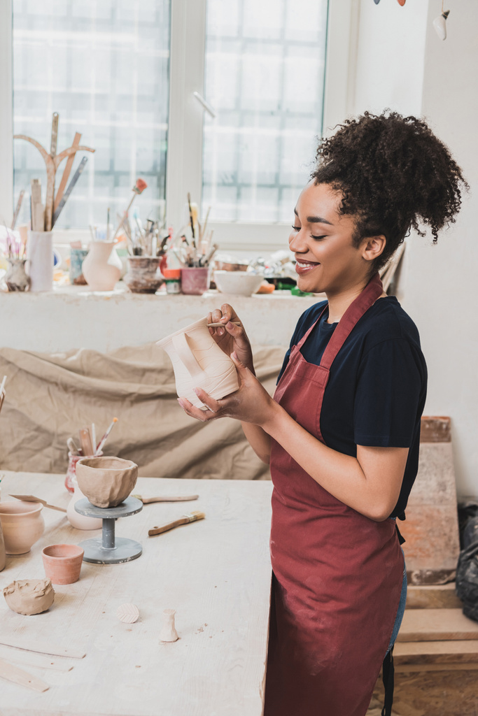 smiling young african american woman sculpting clay pot with stick in pottery - Photo, Image