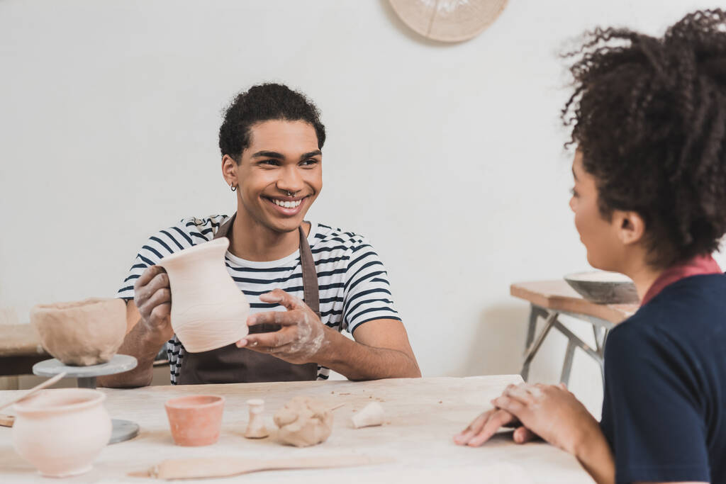 sonriente joven afroamericano hombre mostrando arcilla olla a mujer cerca en cerámica - Foto, imagen
