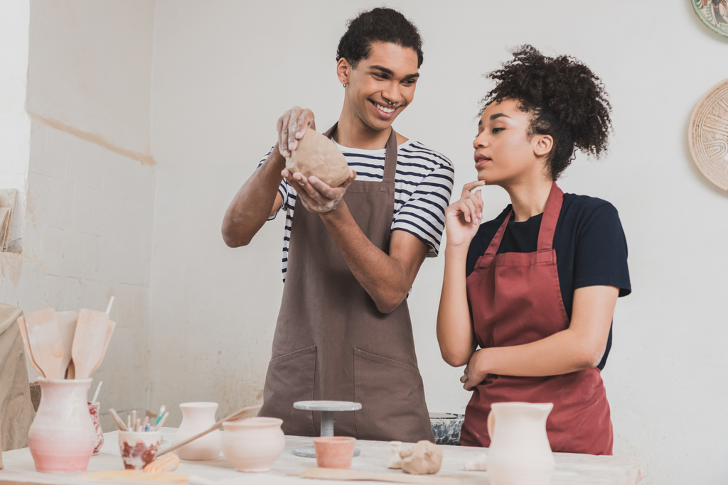 lächelnder junger afrikanisch-amerikanischer Mann, der Tongefäße in der Nähe einer Frau mit der Hand in der Nähe des Gesichts aus Keramik herstellt - Foto, Bild